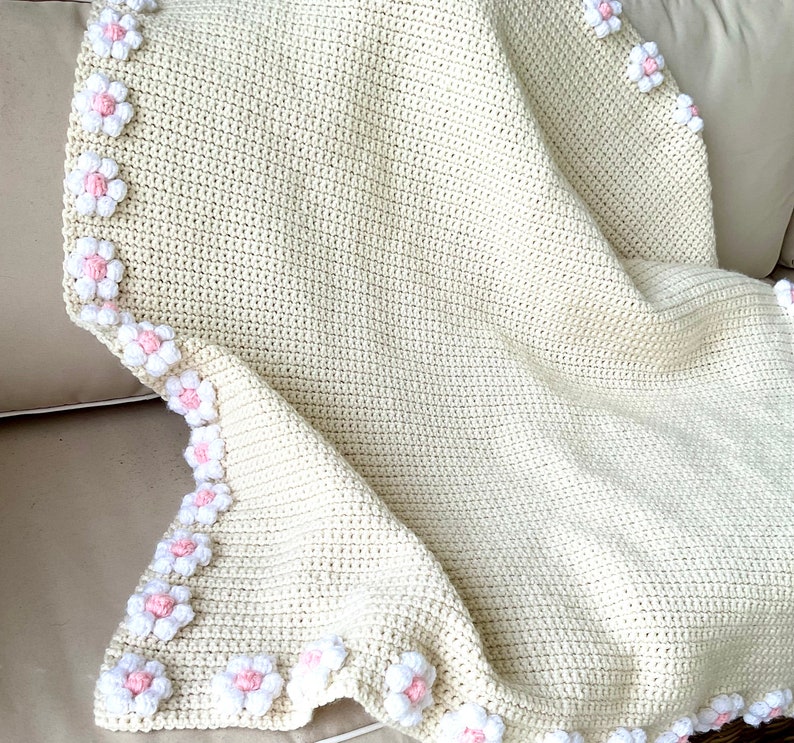 May include: A crocheted baby blanket in cream with a border of pink and white flowers.
