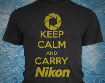 以下が含まれることがあります： 黄色い絞りグラフィックと「KEEP CALM AND CARRY Nikon」の文字が入った濃い灰色のTシャツ。Tシャツはクルーネックで半袖です。テキストとグラフィックはシャツの前面の中央に配置されています。
