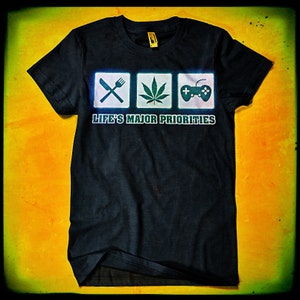 May include: Black t-shirt with a graphic of three icons: a fork and knife, a marijuana leaf, and a video game controller. The text "Life's Major Priorities" is printed below the icons.