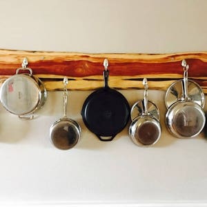 May include: A wooden rack with hooks holding a variety of pots and pans. The rack is made of natural wood with a reddish hue and has a rustic look. The pots and pans are a mix of stainless steel and cast iron.