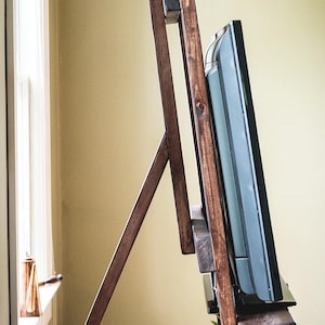 Puede incluir: Un soporte de TV de madera con un tinte marr&oacute;n oscuro. El soporte tiene dos estantes y est&aacute; dise&ntilde;ado para sostener un televisor de pantalla plana. Una planta en maceta se encuentra en el estante inferior.