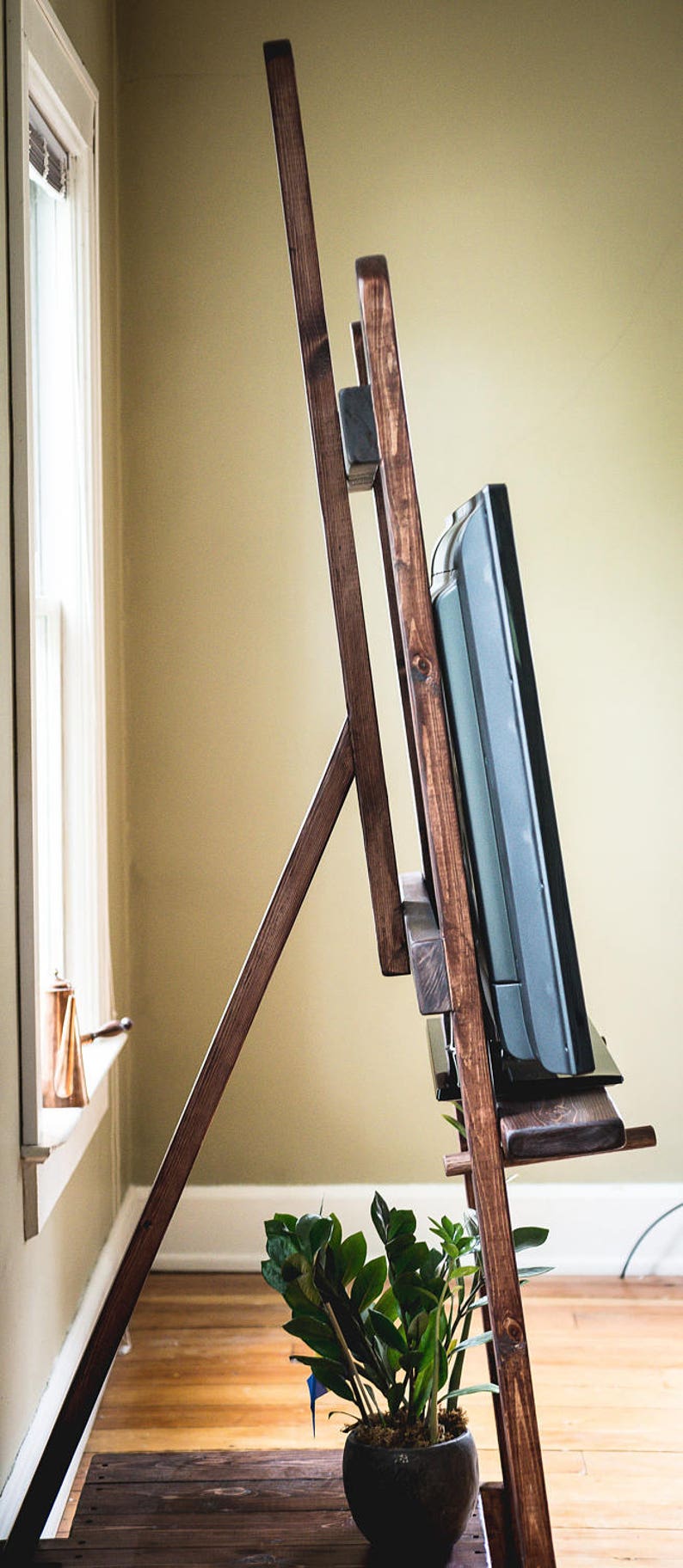 Puede incluir: Un soporte de TV de madera con un tinte marr&oacute;n oscuro. El soporte tiene dos estantes y est&aacute; dise&ntilde;ado para sostener un televisor de pantalla plana. Una planta en maceta se encuentra en el estante inferior.