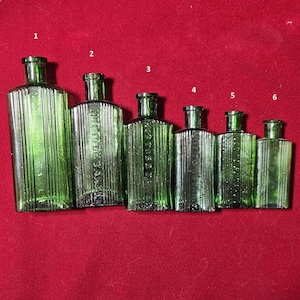 May include: Six antique green glass bottles of varying sizes are arranged on a red surface. The bottles have vertical ribbed detailing and a rectangular shape. Some bottles have text that reads "NOT TO BE TAKEN".