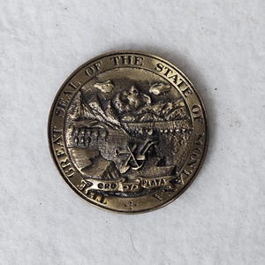 May include: A gold-colored metal coin with the words "The Great Seal of the State of Montana" around the edge. The center of the coin features a depiction of mountains, a river, a plow, a pickaxe, and an anchor.
