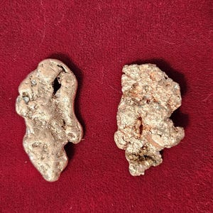 May include: Two irregularly shaped, metallic nuggets with a rough texture. One is a silver color, the other is a bronze color. They are displayed against a deep red background, creating a contrast that highlights their unique forms.