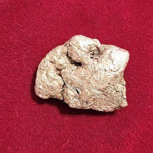 May include: A rough, irregularly shaped, metallic-looking rock with a textured surface. The rock has a light, silvery-gold color and sits on a deep red background.