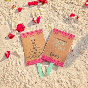 Puede incluir: Dos programas de ceremonia de boda de papel marrón con ribetes de encaje rosa y dorado y mangos de madera turquesa. Los programas están en una playa de arena blanca con pétalos de rosa rosa esparcidos alrededor. El texto en los programas dice "The Wedding Ceremony of Keisha & Mark" y "August 2nd, 2018, Destin, Florida".