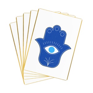 Può includere: Un mazzo di carte bianche con bordi dorati, ciascuna con una mano di hamsa blu con un occhio turco blu chiaro al centro. La mano di hamsa è un simbolo di protezione e buona fortuna.