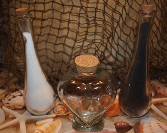 May include: Two tall, slender glass bottles with cork stoppers, one filled with white sand and the other with black sand, positioned on either side of a central heart-shaped glass bottle with a cork stopper.