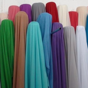 May include: A collection of fabric bolts displayed in a vertical arrangement, featuring a wide spectrum of solid colors including teal, lavender, green, tan, turquoise, blue, purple, gray, white, red, and dark maroon, creating a vibrant display.