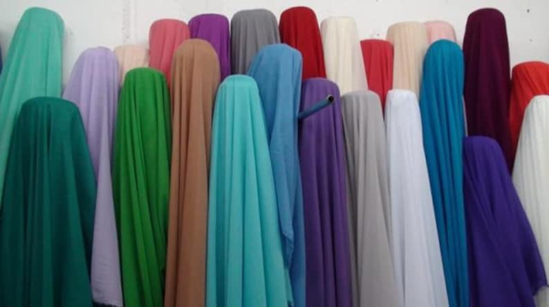 May include: A collection of fabric bolts displayed in a vertical arrangement, featuring a wide spectrum of solid colors including teal, lavender, green, tan, turquoise, blue, purple, gray, white, red, and dark maroon, creating a vibrant display.