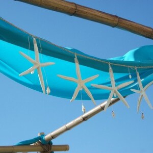 May include: A decorative garland featuring white starfish and small seashells suspended from a string, draped against a bright turquoise fabric canopy. The arrangement is supported by horizontal bamboo poles against a clear blue sky background.