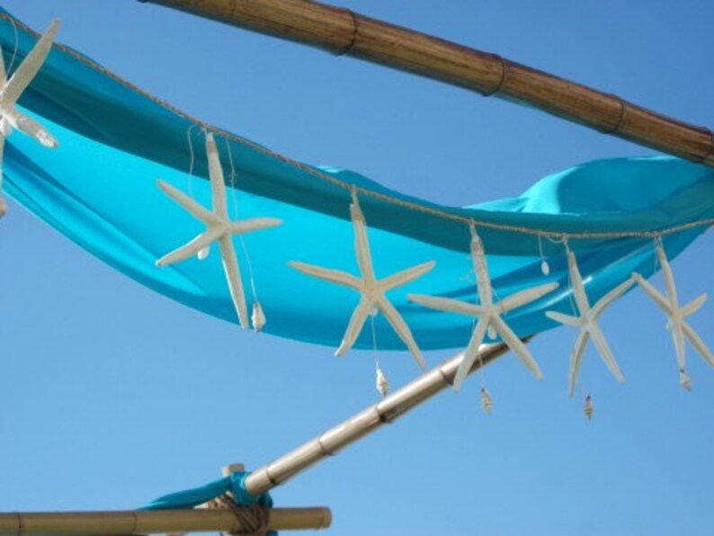 May include: A decorative garland featuring white starfish and small seashells suspended from a string, draped against a bright turquoise fabric canopy. The arrangement is supported by horizontal bamboo poles against a clear blue sky background.