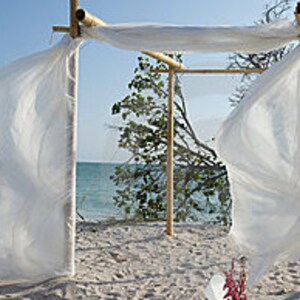 May include: A bamboo frame structure featuring four vertical poles and horizontal crossbeams, draped with long, flowing white fabric panels that hang from the top and sides, creating a rectangular archway design for an outdoor setting.