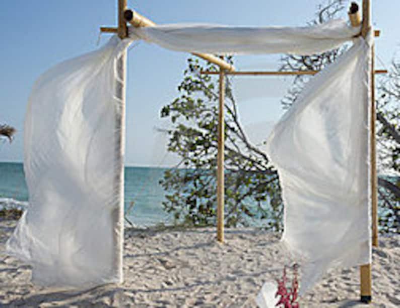 May include: A bamboo frame structure featuring four vertical poles and horizontal crossbeams, draped with long, flowing white fabric panels that hang from the top and sides, creating a rectangular archway design for an outdoor setting.