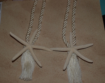 May include: Two off-white starfish curtain tiebacks featuring twisted rope cords and matching tassels. Each starfish is attached to a decorative cord, providing a coastal aesthetic for window treatments. The set includes two identical pieces.