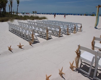 May include: A beach wedding setup with rows of white chairs on the sand. Each chair is decorated with starfish and twine. The ceremony backdrop is decorated with flowing fabric. The beach is sunny with blue skies.