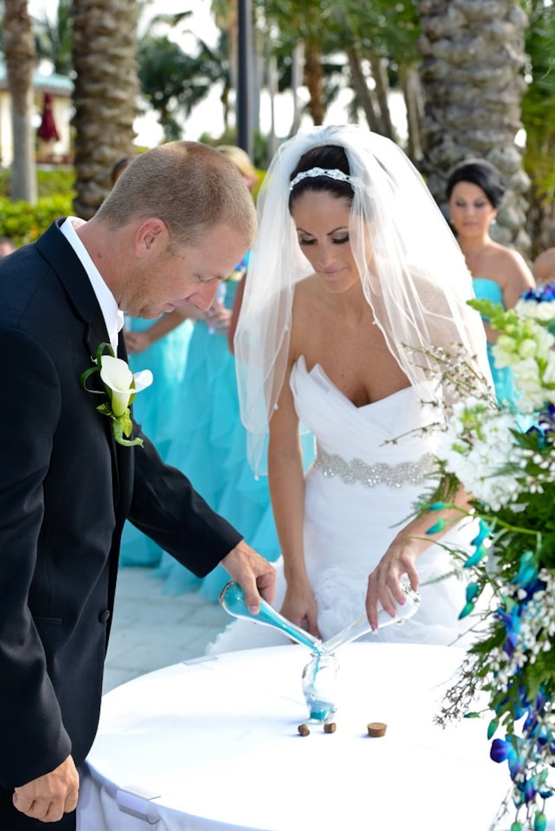 May include: A bride and groom pour blue sand into a glass vase during a wedding ceremony. The bride is wearing a white wedding dress with a veil and the groom is wearing a black suit. The ceremony is taking place outdoors with palm trees in the background.