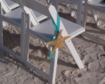 May include: White folding chairs decorated with a turquoise ribbon and a starfish on a sandy beach.