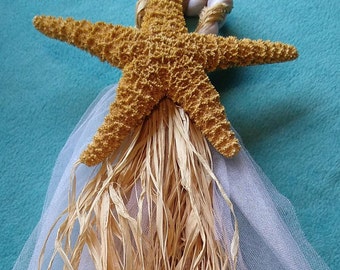 May include: A decorative starfish ornament with a natural, textured appearance. It features a woven rope loop for hanging, a white tulle skirt, and dried grass accents. The starfish is a warm, sandy brown color.