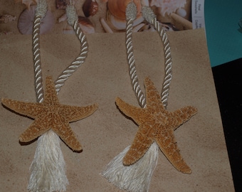 May include: A pair of curtain tiebacks featuring natural tan starfish attached to cream-colored twisted rope cords. Each tieback includes a decorative cream-colored tassel hanging from the base of the starfish, providing a textured nautical accent.