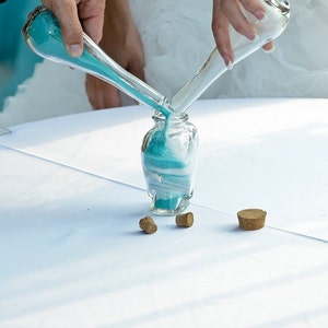 May include: Two clear glass bottles with blue and white sand are being poured into a larger clear glass bottle. The bottles are on a white tablecloth. There are three brown bottle stoppers on the table.