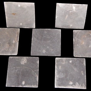 May include: Seven square, translucent, off-white tiles with a marbled texture. Each tile features two small, circular holes positioned near the edges. The tiles are arranged in a cluster against a solid black background, showing natural variations.