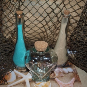 May include: Three glass bottles with cork stoppers, two are filled with blue and white sand, the third is empty and heart-shaped. The bottles are sitting on a brown surface with a net background.