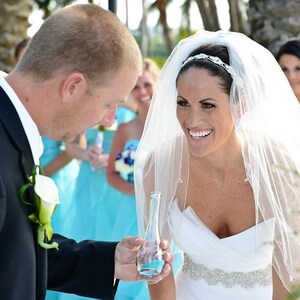 May include: A bride in a white wedding dress with a veil smiles as she holds a small glass bottle. The groom in a black suit holds the bottle with her. The bride is wearing a silver headband and a silver belt. The bridesmaids are wearing turquoise dresses.