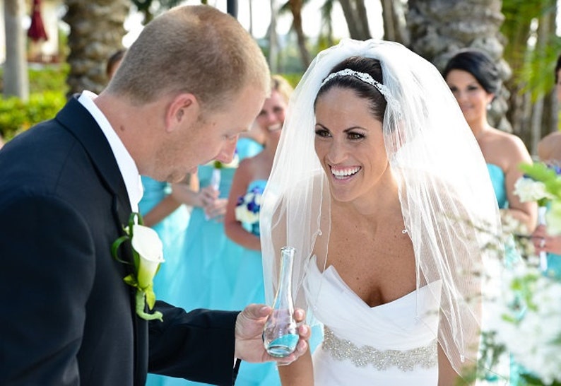 May include: A bride in a white wedding dress with a veil smiles as she holds a small glass bottle. The groom in a black suit holds the bottle with her. The bride is wearing a silver headband and a silver belt. The bridesmaids are wearing turquoise dresses.