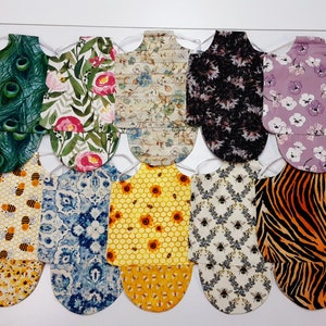 May include: A collection of fabric bibs with various patterns, including peacock feathers, floral designs, bees, sunflowers, and animal prints. Each bib is attached to a white hanger, showcasing the different designs and colors.