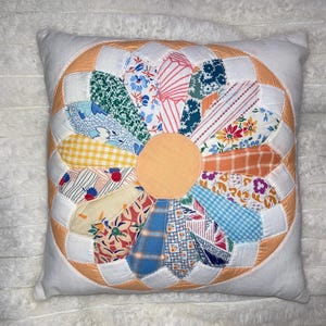May include: Square white pillow with a large, colorful flower design. The flower has a peach-colored center and petals made of various patterned fabrics in shades of blue, yellow, orange, and pink. The flower is surrounded by a peach-colored ring.