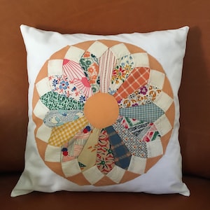 May include: A white pillow with a patchwork flower design in shades of orange, pink, yellow, blue, and green. The flower is made of various fabrics with floral and geometric patterns.