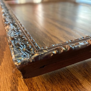 May include: Close-up of a dark brown, ornate picture frame with detailed carvings. The frame has a distressed finish with hints of gold and is set against a wooden surface. The frame's corner is visible.
