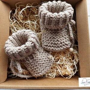 Könnte beinhalten: Zwei Paar beige gestrickte Babyschuhe mit Holzperlen, verpackt in einem braunen Karton mit zerkleinertem Papier.