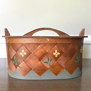 May include: An oval wooden basket with a woven lattice pattern, featuring a central arched handle and two side handles. The exterior is decorated with hand-painted yellow flowers and green leaves, with a solid grey painted base border.