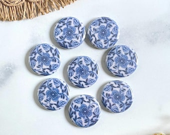 May include: Seven circular buttons featuring a white base with a blue floral pattern. Each button displays a central flower surrounded by intricate leafy vines and petals, arranged in a cluster on a smooth, light-colored surface.