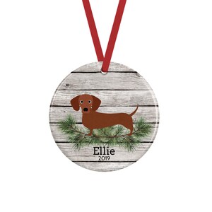 May include: A round ceramic ornament featuring a brown dachshund standing on green pine branches against a light gray wood-plank background. The name Ellie and the year 2019 are printed in black text below the dog. A red ribbon is attached to the top.