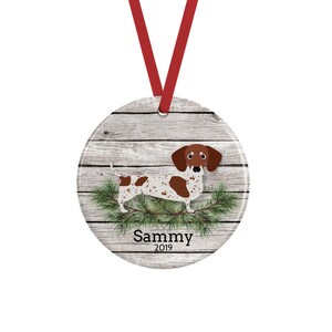May include: A round ceramic ornament featuring a brown and white spotted dachshund standing on green pine branches against a gray wood plank background. The name Sammy and the year 2019 are printed in black text below the dog. A red ribbon is attached.
