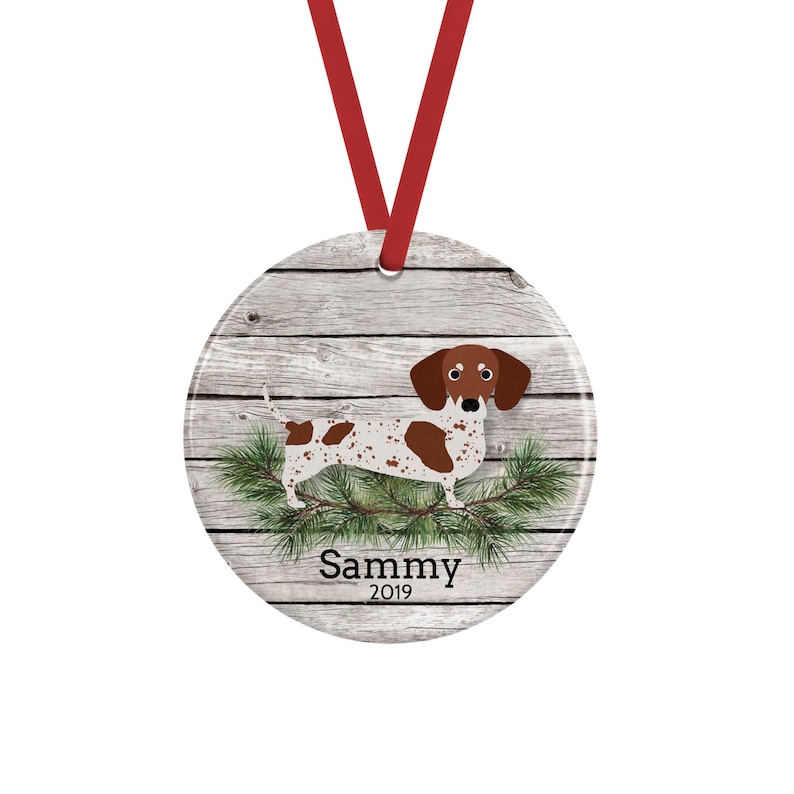 May include: A round ceramic ornament featuring a brown and white spotted dachshund standing on green pine branches against a gray wood plank background. The name Sammy and the year 2019 are printed in black text below the dog. A red ribbon is attached.