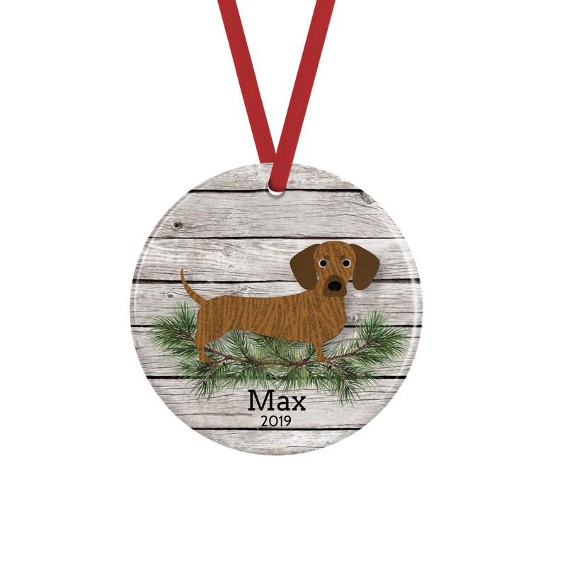 May include: A round ceramic ornament featuring a brown dachshund standing on green pine needles against a light gray wood grain background. The text Max 2019 is printed in black below the dog. A red ribbon is attached to the top for hanging.