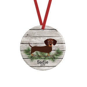 May include: A round ceramic ornament featuring a brown dachshund standing on green pine branches against a light gray wood grain background. The name Sadie and the year 2019 are printed in black text below the dog. A red ribbon is attached for hanging.