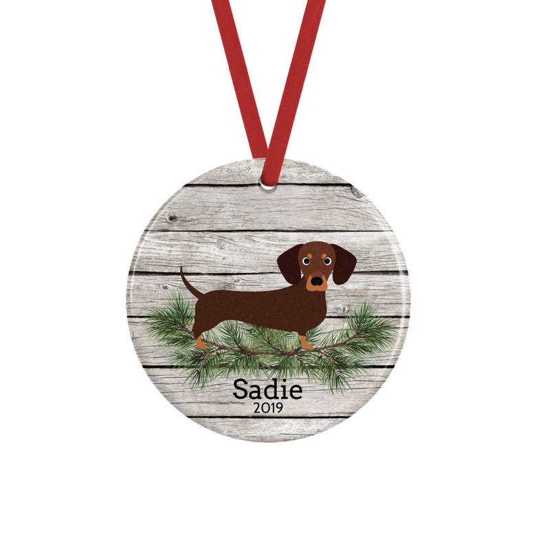 May include: A round ceramic ornament featuring a brown dachshund standing on green pine branches against a light gray wood grain background. The name Sadie and the year 2019 are printed in black text below the dog. A red ribbon is attached for hanging.