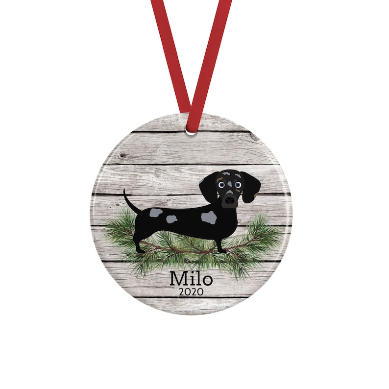 May include: A round ceramic ornament featuring a black and white dachshund illustration standing on green pine needles against a gray wood-plank background. The text Milo and 2020 is printed in black below the dog. A red ribbon is attached to the top.