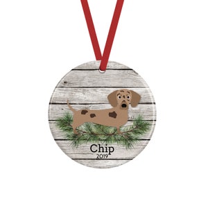 May include: A round ceramic ornament featuring a light brown dachshund with dark brown spots, standing on green pine needles against a light gray wood grain background. The text Chip and 2019 is printed in black below the dog, with a red ribbon.