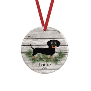 May include: A round ceramic ornament hanging from a red ribbon, featuring a black and tan dachshund illustration standing on green pine branches against a gray wood-plank background, with the text Louie and the year 2019 printed in black.