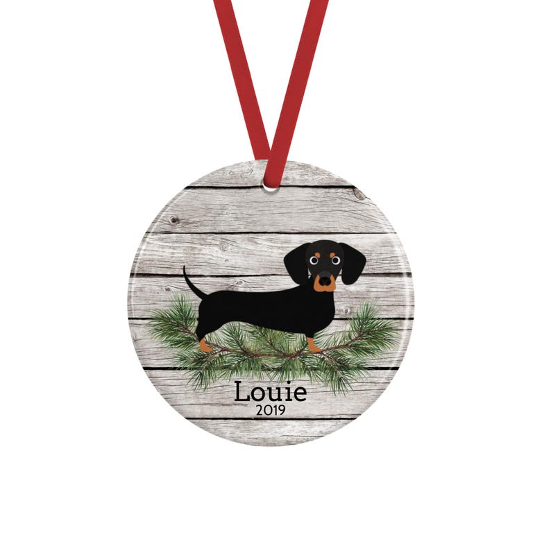 May include: A round ceramic ornament hanging from a red ribbon, featuring a black and tan dachshund illustration standing on green pine branches against a gray wood-plank background, with the text Louie and the year 2019 printed in black.