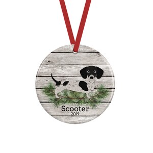May include: A round ceramic ornament featuring a black and white spotted dachshund illustration over a gray wood grain background with green pine needles. The text Scooter and 2019 are printed below the dog. A red ribbon is attached for hanging.