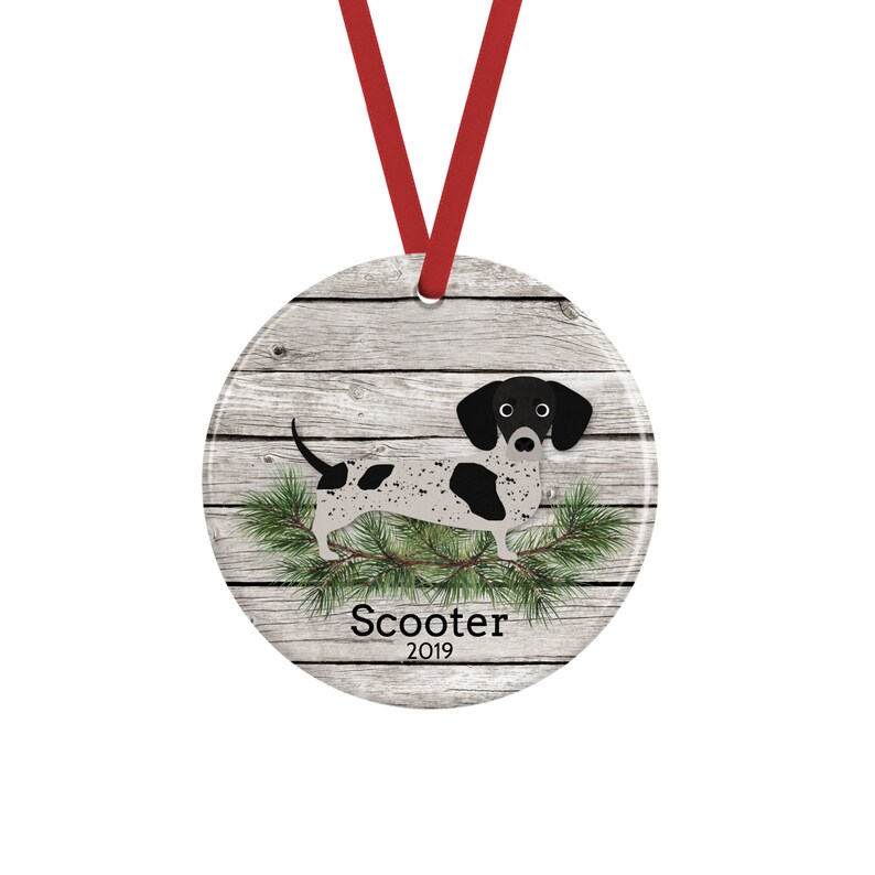 May include: A round ceramic ornament featuring a black and white spotted dachshund illustration over a gray wood grain background with green pine needles. The text Scooter and 2019 are printed below the dog. A red ribbon is attached for hanging.