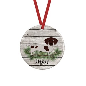 May include: A round ceramic ornament featuring a brown and white dachshund illustration over a gray wood grain background with green pine needles. The text Henry 2019 is printed in black below the dog. A red ribbon is attached for hanging.